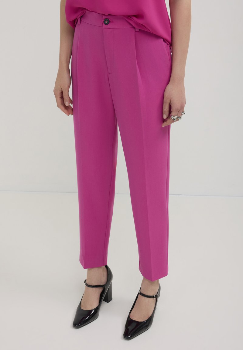 Pantaloni fucsia con vestibilità sartoriale, dotati di chiusura a bottoni e pieghe, abbinati a scarpe nere con tacco e una blusa coordinata.