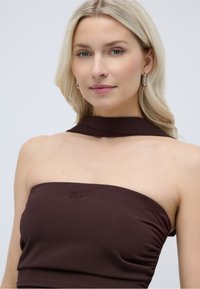 Ξανθιά γυναίκα που φορά ασημένια σκουλαρίκια κρίκους και ένα σκουρό καφέ τοπ χωρίς τιράντες με λεπτομέρεια στο λαιμό που ταιριάζει, μπροστά σε ένα απλό ανοιχτόχρωμο φόντο.