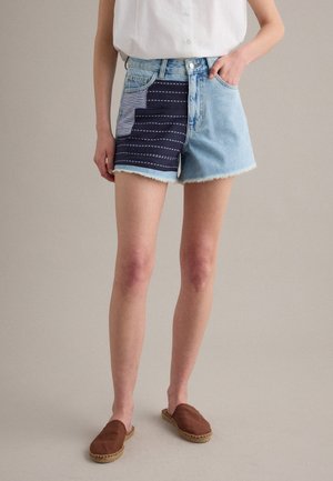 Femme portant un short en denim avec une pièce bleu foncé et un motif à rayures blanches, un chemisier blanc sans manches et des chaussures marron sans lacets.