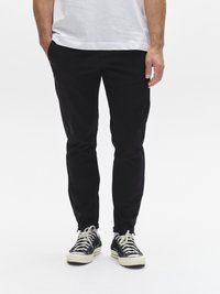 Melnas, plānas bikses no vieglas auduma, ar sānu kabatām un rāvējslēdzēja aizdari pie bikšu galiem, kopā ar melniem un baltiem sporta apaviem.