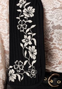 Zwarte stof met witte bloemrijke borduurwerk, voorzien van een doorgestikt ontwerp en een gebogen vorm. Op de achtergrond is een contrasterende kanttextuur zichtbaar.