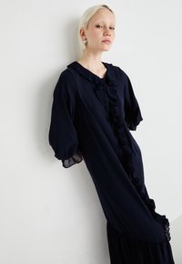 Vestido azul marinho com colarinho volumoso e detalhe frontal. Apresenta mangas balão e uma silhueta fluida com pormenores transparentes.