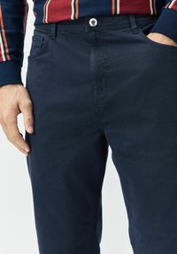 Mann in marineblauen Hosen mit Knopfverschluss und Hand in der rechten Tasche, kombiniert mit einem gestreiften Langarmhemd in Marineblau, Rot und Weiß.