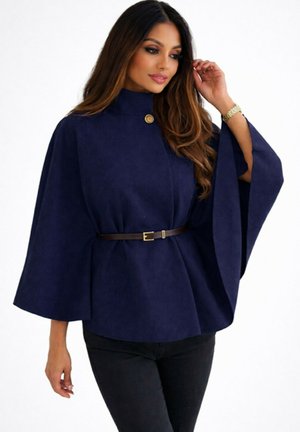 Femme aux longs cheveux portant une cape bleu marine à col montant, avec un bouton et une ceinture, associée à un pantalon noir, posant avec une main près des cheveux.