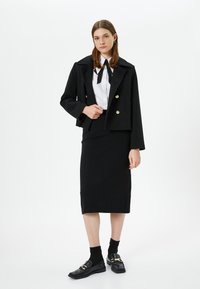 Jonge vrouw in een zwarte cropped blazer, witte blouse met zwarte strik, zwarte midi rok, zwarte sokken en zwarte instappers, staande voor een witte achtergrond.
