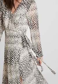 Langes Wickelkleid aus einem durchsichtigen, leichten Stoff mit beigem Grund und schwarzen sowie braunen Tiermustern. Mit Bändern in der Taille und langen Ärmeln.
