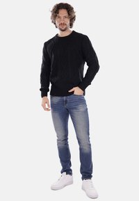 Maglione nero a maglia con design a trecce, polsini a costine, indossato sopra jeans blu e sneakers bianche. Il modello si trova in piedi con una mano in tasca.