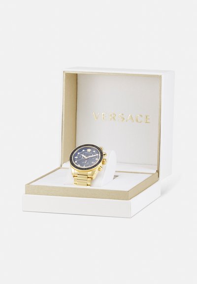 Kuldne käekell musta dialsiga, kuldse tooniga rihm ja hõbedased aksessuaarsed, eksponeeritud valges ja kuldse karbis, millel on "Versace" logo.
