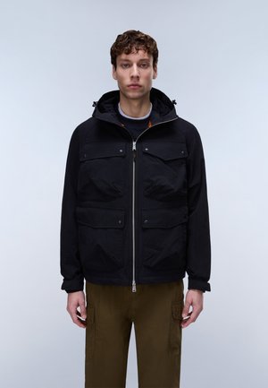 Ung mand med krøllet hår iført en sort hættetrøje med flere lommer og olivengrønne cargobukser, stående mod en neutral baggrund.