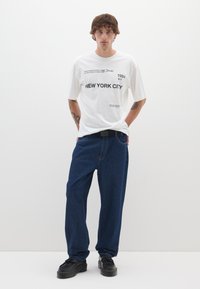 Camiseta blanca con gráficos negros que dicen "NEW YORK CITY" y "1984 NY". Pantalones vaqueros de denim azul oscuro de corte holgado. Zapatos negros con suelas gruesas.