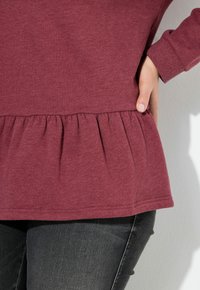 Top burdeos con mangas largas y dobladillo peplum fruncido, fabricado en tela suave. Detalles de textura visibles en la cintura y las mangas.
