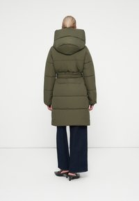 Olive green padded coat with a large hood, cinched waist, and long sleeves, paired with wide-leg jeans and black heels.