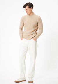 Beige stickad tröja med hög krage och flätmönster, ihop med vita byxor och ljusa sneakers. Modellen står med händerna i fickorna.