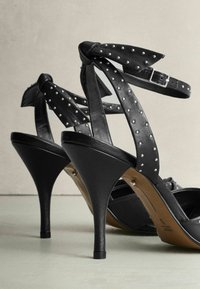 Schwarze High-Heelsandalen mit genoppten Riemen, ausgestattet mit einem Schleifendetail, aus glattem Leder und mit einem eleganten, spitz zulaufenden Design.