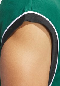 Zelené sportovní tričko z látky s černým zakončením v podpaží a bílým akcentem na výstřihu. Detailní záběr na rameno odhaluje hladkou texturu pleti.