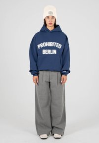Sudadera azul marino con "PROHIBIDO BERLÍN" en letras blancas, combinada con pantalones gris de pierna ancha. Los accesorios incluyen un gorro de punto crema.