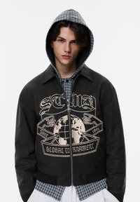 Musta bomber-takki vetoketjulla, jossa on maailmankilpailua kuvaava kermaan taittuva grafiikka ja kontrastina ruudullinen hupun vuori.