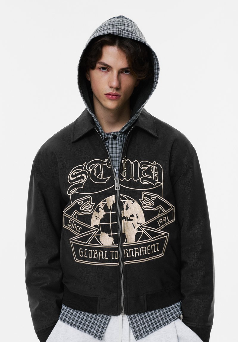 Schwarze Bomberjacke mit Reißverschluss, versehen mit einem globalen Turniergrafik in Cremefarbe und einem karierten Kapuzenfutter als Kontrast.