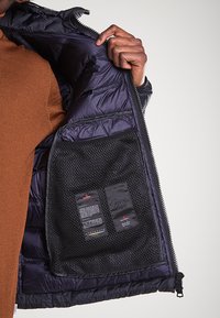 Giubbotto isolato blu scuro con un design a trapunta, dotato di una tasca interna in rete e etichette per dettagli di cura e materiale.