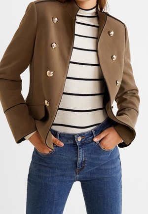 Veste marron croisée avec des boutons dorés sur une chemise rayée blanche et noire, associée à un jean bleu et les mains dans les poches.