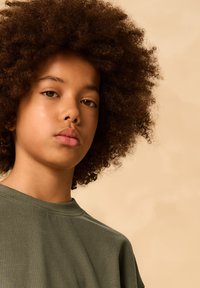 Tricou verde din bumbac, cu decolteu rotund, croială ușor largă și mâneci scurte, purtat de o persoană cu păr creț și maro, pe un fundal neutru.