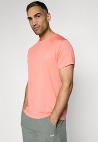 Tricou scurt cu mâneci din material texturat, de culoare corai, cu decolteu rotund și un logo mic pe piept, asortat cu pantaloni scurți gri deschis.