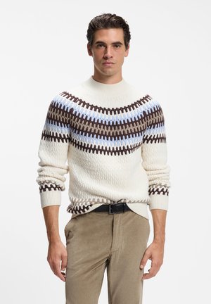 Homme portant un pull crème avec empiècement marron et bleu à motifs et un pantalon en velours côtelé beige, debout devant un fond blanc.