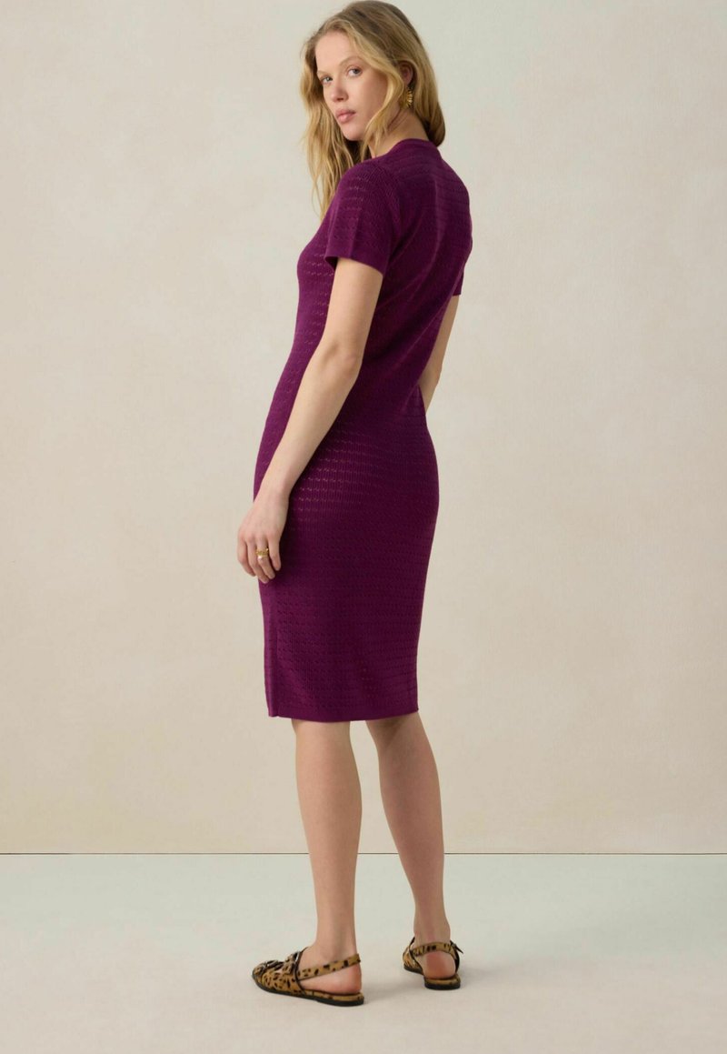 Enganliegendes lila Strickkleid mit kurzen Ärmeln, strukturiertem Muster und knielanger Schnittführung. Kombiniert mit Tierfell-Print Sandalen. Neutraler Hintergrund.