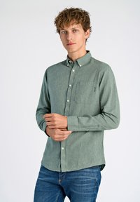Camisa de botón verde de manga larga con un bolsillo, hecha de una tela texturizada. Cuenta con un collar clásico y puños abotonados, combinada con jeans azules.