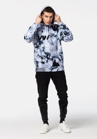 Sudadera con capucha en azul con estampado tie-dye y patrones abstractos en negro. Cuenta con un bolsillo canguro, cordones blancos y puños acanalados. Pantalones y zapatos negros.