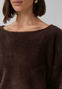 Brązowy, puszysty sweter dzianinowy z szerokim dekoltem. Posiada miękką teksturę i luźny krój, podkreślający sylwetkę odkrywającą ramię.