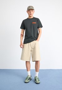 Black t-shirt with red "OBEY" logo, beige plaid shorts, green sneakers, and a matching cap. Model poses on a blue carpeted surface.