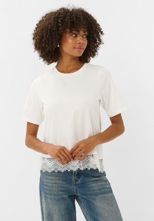 Femme aux cheveux bouclés portant un haut blanc à manches courtes avec bordure en dentelle et un jean bleu, regardant légèrement sur le côté avec les mains jointes.