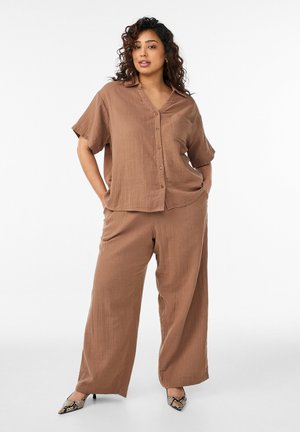 Kobieta o kręconych włosach ubrana w luźną brązową koszulę na guziki i pasujące szerokie spodnie, stoi z rękami w kieszeniach na neutralnym tle.