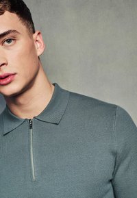 Světle zelené polo tričko na zip s texturovanou tkaninou, s límcem a polo-zip uzavřením, vystavené na osobě na neutrálním pozadí.