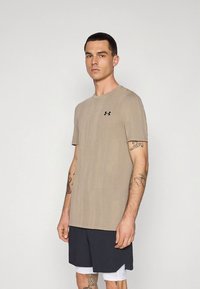 Béžové tričko s krátkým rukávem z měkké látky, s kulatým výstřihem a malým černým logem na hrudi. Hladká textura.