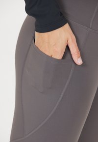 Grå leggings lavet af elastisk materiale, med en sidelomme og flade sømme for komfort. Nærbillede viser lommedetaljen.