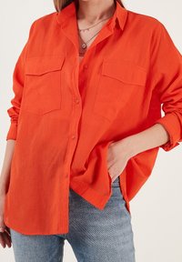Femme portant une chemise orange vif à boutons avec les manches repliées, un jean en denim clair, et des colliers délicats superposés, main dans la poche.