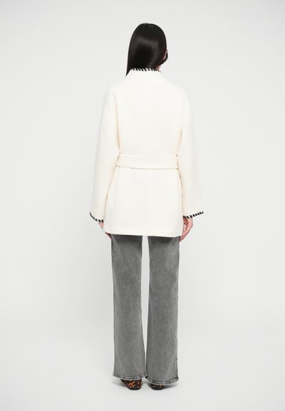 Manteau beige, en mélange de laine, avec une taille ceinturée, présentant des finitions noires au col et aux manches ; associé à un pantalon large gris.