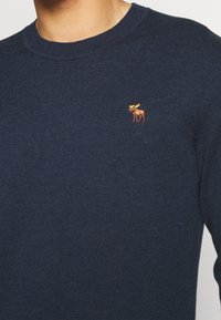 Navyblauer Strickpullover mit runder Halslinie und einem kleinen gestickten Elch-Logo in Orange und Gelb auf der Brust.