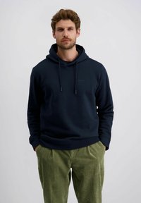 Marineblaue Kapuzenjacke mit Kordelzug und einem minimalistischen Logo; kombiniert mit strukturierten olivgrünen Cordhosen.