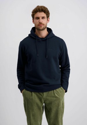 Marineblaue Kapuzenjacke mit Kordelzug und einem minimalistischen Logo; kombiniert mit strukturierten olivgrünen Cordhosen.