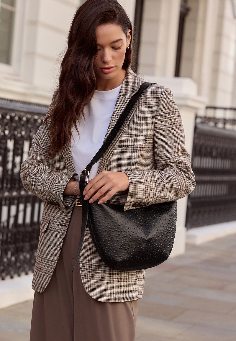 Woman wearing checked blazer and white shirt holding a textured black shoulder bag on a city sidewalk.