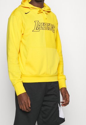 Mand iført en lys gul Lakers-hættetrøje med frontlomme og sort Nike-logo, kombineret med sorte sportsshorts med hvide detaljer.