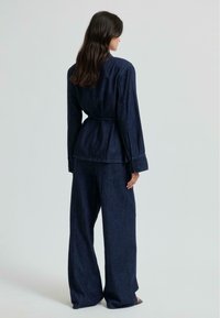 Temno modra denim jakna s pritegnjenim pasom, v kombinaciji s širokimi temno modrimi denim hlačami. Ima dolge rokave in minimalističen dizajn.