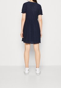 Marinblå bomulls klänning med ögla detaljer, korta volangärmar och figurnära midja. Matcher med vita högtopps sneakers.