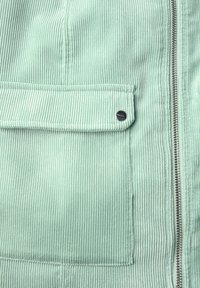 Hellgrüne Cordjacke mit vertikal gerippter Textur, Reißverschluss und einer aufgesetzten Tasche mit einem runden schwarzen Knopf.