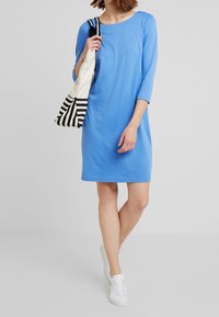 Blauwe katoenen jurk met driekwartmouwen, ronde hals, rechte snit, gecombineerd met een gestreepte tote bag en witte sneakers.