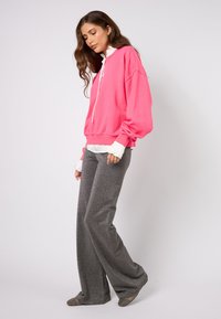 Svetlo ružová nadrozmerná mikina s bielymi šnúrkami, navrchu biela blúzka s volánikmi, v kombinácii s trblietavými sivými nohavicami so širokými nohavicami.