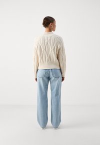 Πλεκτό πουλόβερ σε κρέμα με γεωμετρικά υφασματικά σχέδια, συνδυασμένο με ανοιχτό μπλε παντελόνια φαρδιάς γραμμής και λευκά αθλητικά παπούτσια. Όψη από πίσω.